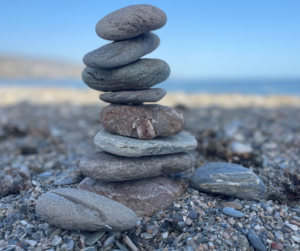in het midden van de afbeelding staat een stapel van 8 gladde afgeronde stenen. Ze staan op een stenenstrand met een blauwe lucht in de achtergrond