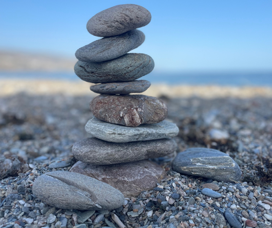 in het midden van de afbeelding staat een stapel van 8 gladde afgeronde stenen. Ze staan op een stenenstrand met een blauwe lucht in de achtergrond
