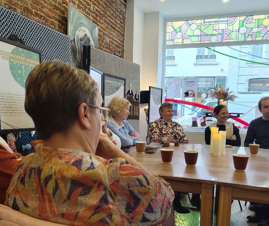 een groep van 7 mensen zit om een tafel met koppen koffie erop en zijn met elkaar in gesprek