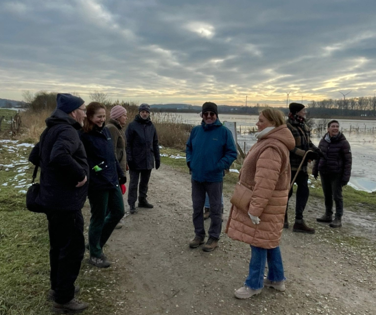 een groep van 8 wandelaars staat op een zanderig wandelpad naast een poel met water. Ze dragen allemaal warme kleding, mutsen en sjaals. Het meer is bevroren en langs de kanten van het wandelpad ligt sneeuw.