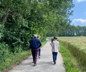 4 wandelaars lopen over een bestraat wandelpad. Links van het pad staan bomen en struiken, rechts is een weiland met hoog gras. De zon schijnt en de lucht is blauw.