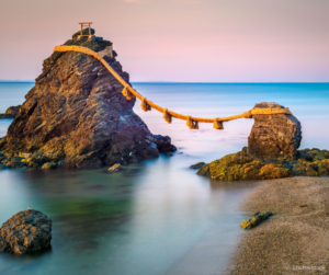 een kalme zee met een paarse lucht op de achtergrond. Op de voorgrond staan twee rotsen, op de grotere rots aan de linkerkant staat een kleine shinto tempel. De rots is met een touw met decoratieve kwasten verbonden aan de kleine rots aan de rechterkant.