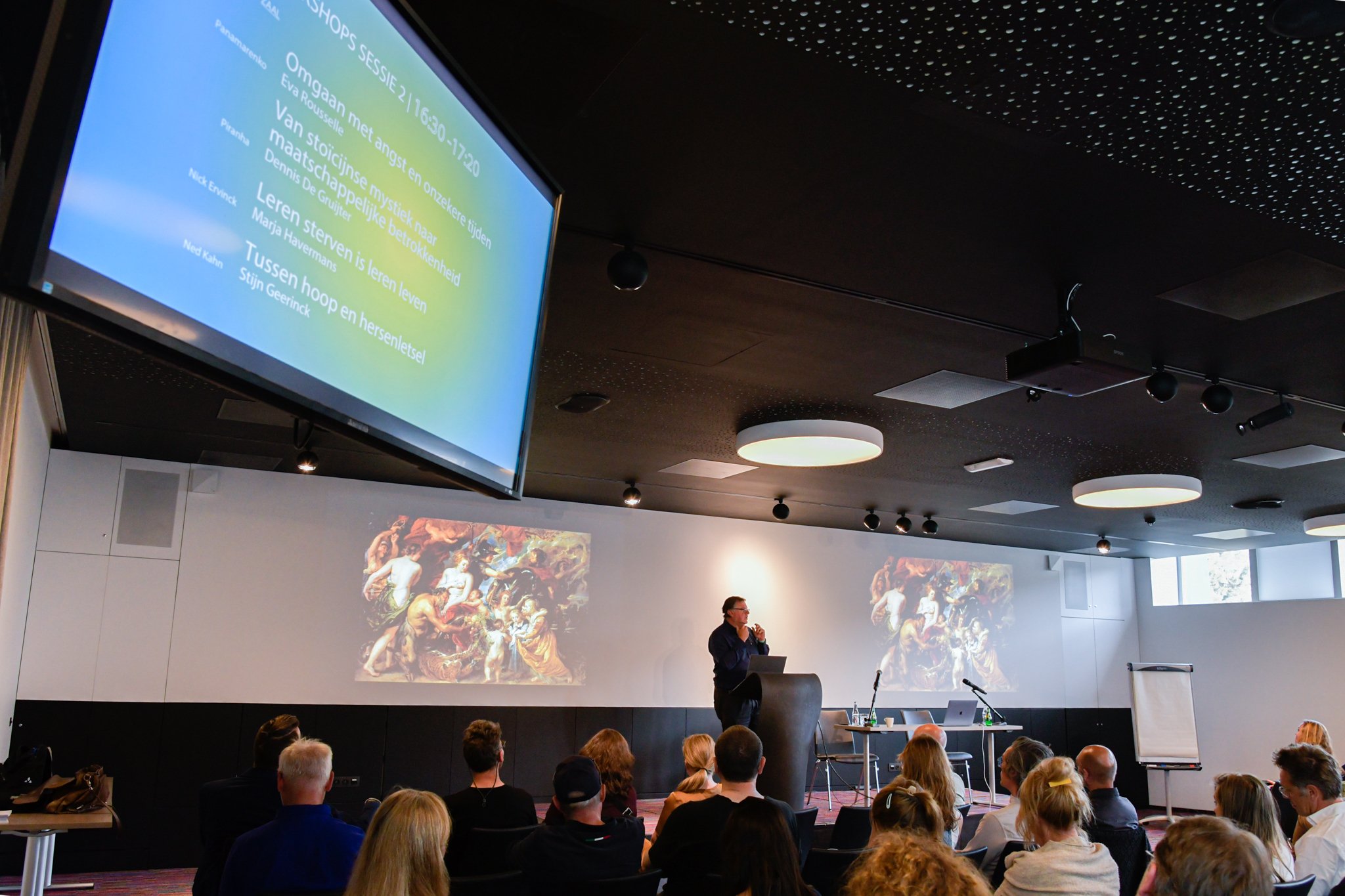 Een spreker staat op een podium voor een zaal met aanwezigen in een moderne conferentieruimte. Achter de spreker worden twee grote projecties getoond: een dia met tekst en een afbeelding van een klassiek schilderij. Rechts staat een flip-over en op de tafel naast de spreker liggen enkele materialen en een microfoon