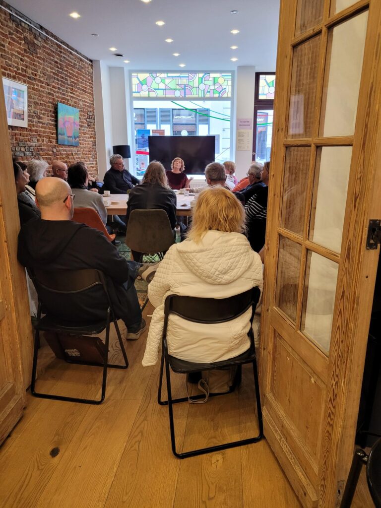 een groep oudere mensen zit rondom een tafel, ze luisteren allemaal aandachtig naar de vrouw die spreekt