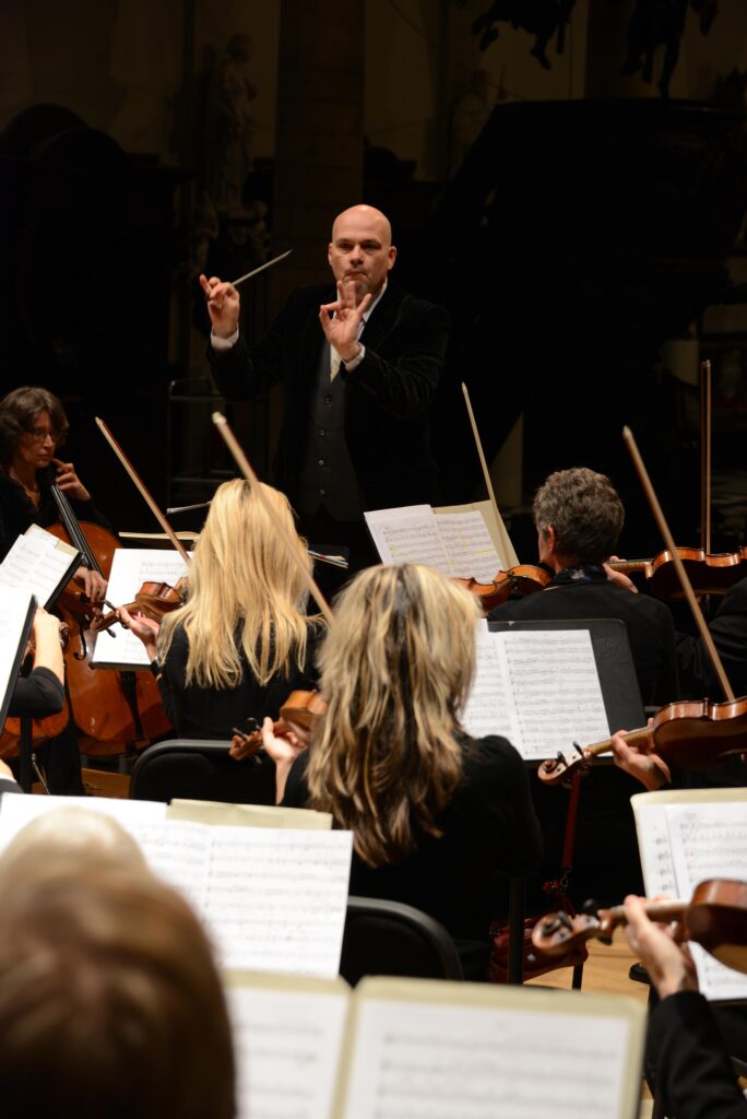 dirigent rik ghesquiere van het belle epoque orchestra staat op de bok met zijn gezicht naar de camera. Voor hem zitten meerdere violisten met hun rug naar de camera terwijl zij hun instrument bespelen
