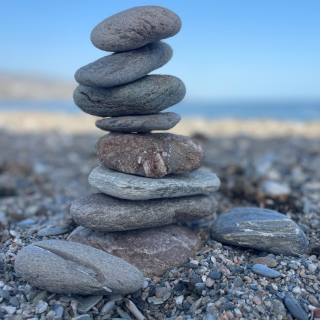 in het midden van de afbeelding staat een stapel van 8 gladde afgeronde stenen. Ze staan op een stenenstrand met een blauwe lucht in de achtergrond