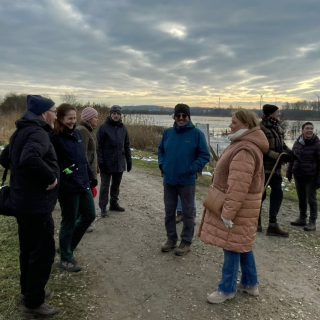 een groep van 8 wandelaars staat op een zanderig wandelpad naast een poel met water. Ze dragen allemaal warme kleding, mutsen en sjaals. Het meer is bevroren en langs de kanten van het wandelpad ligt sneeuw.