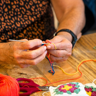 Haakcafe met een tafel met bollen wol in verschillende kleuren. aan tafel zitten een vrouw met een haaknaald het begin van een knuffel te maken