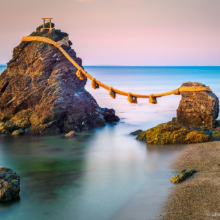 een kalme zee met een paarse lucht op de achtergrond. Op de voorgrond staan twee rotsen, op de grotere rots aan de linkerkant staat een kleine shinto tempel. De rots is met een touw met decoratieve kwasten verbonden aan de kleine rots aan de rechterkant.