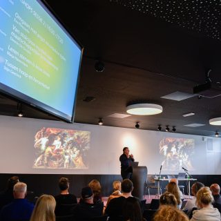 Een spreker staat op een podium voor een zaal met aanwezigen in een moderne conferentieruimte. Achter de spreker worden twee grote projecties getoond: een dia met tekst en een afbeelding van een klassiek schilderij. Rechts staat een flip-over en op de tafel naast de spreker liggen enkele materialen en een microfoon