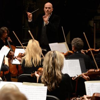 dirigent rik ghesquiere van het belle epoque orchestra staat op de bok met zijn gezicht naar de camera. Voor hem zitten meerdere violisten met hun rug naar de camera terwijl zij hun instrument bespelen